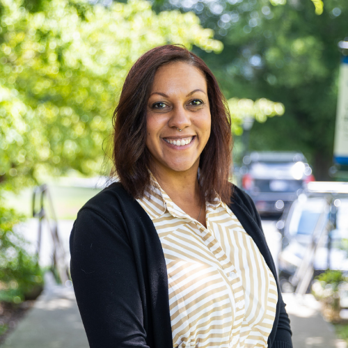 Melanie Bush smiling outdoors, wearing a tan-and-white horizontally striped button-up shirt under a black cardigan, with blurred trees and vehicles in the background.