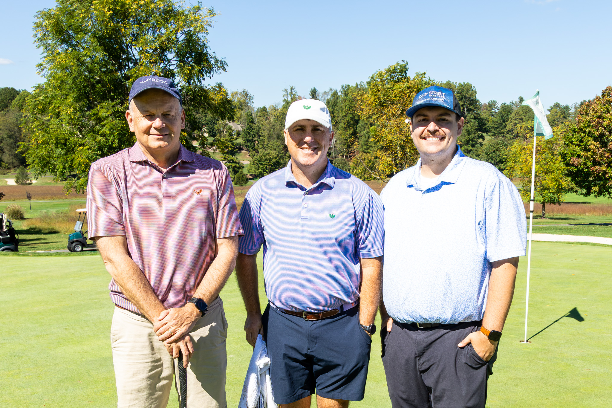 Three male golfers standing together on a golf green during a clear, sunny day, with golf carts, trees, and a flagstick visible in the background.