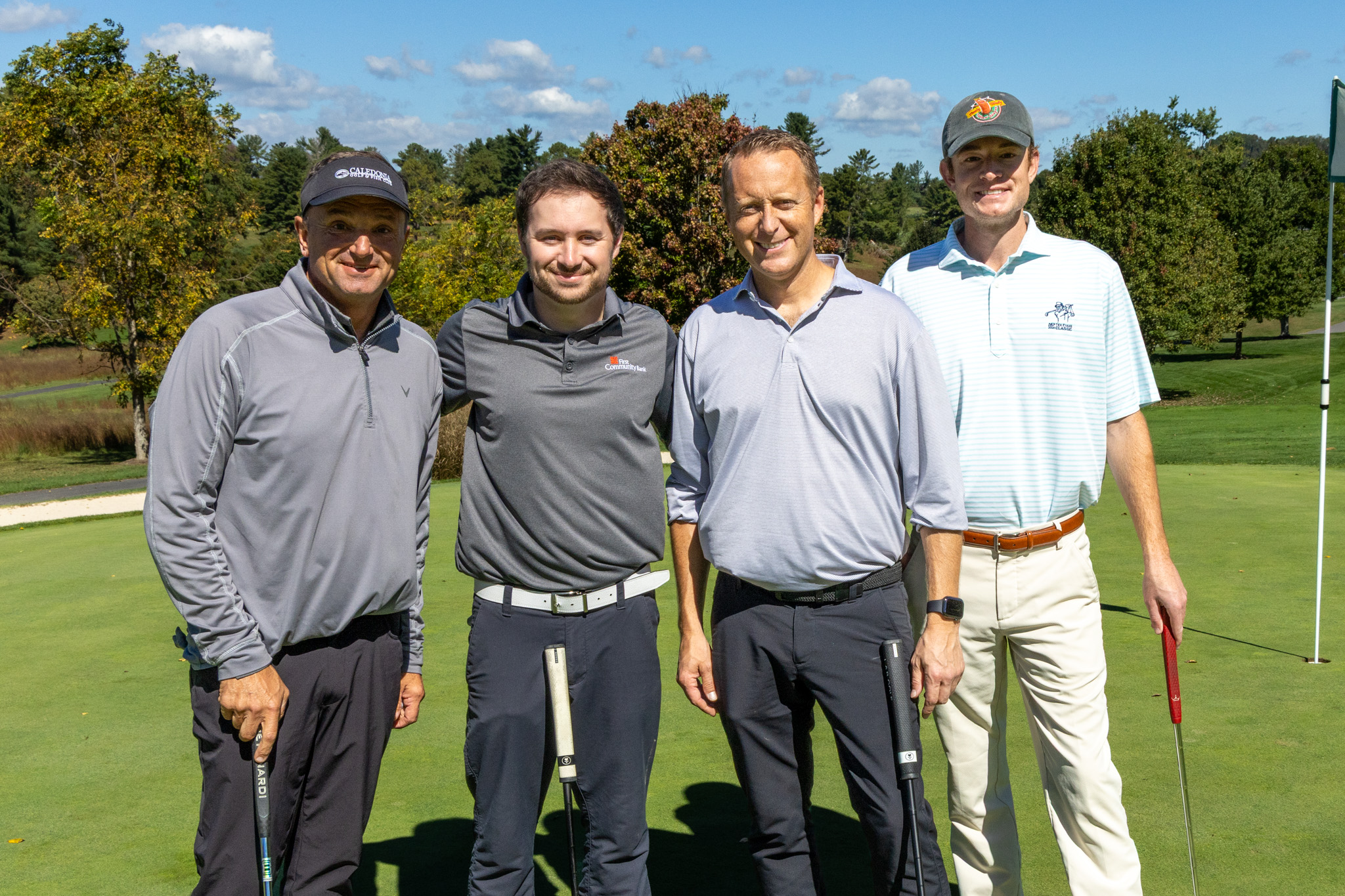Four male golfers smiling while standing together on a golf green, wearing casual golf attire and baseball hats or visors, with a golf flag visible in the background.