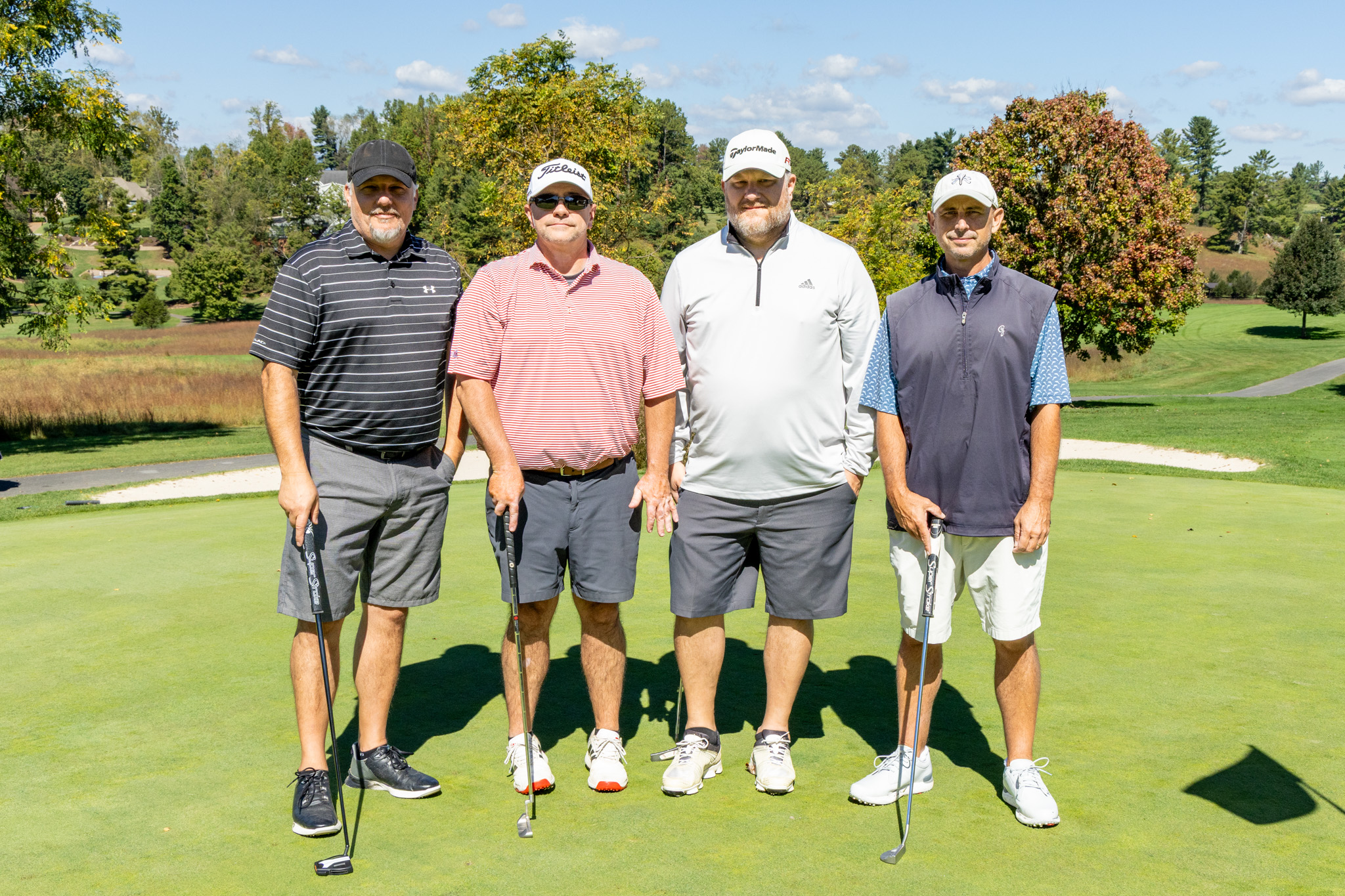 Four male golfers standing side-by-side in a line on a sunny golf green, holding putters and posing for a group portrait with trees and a golf course landscape in the background.
