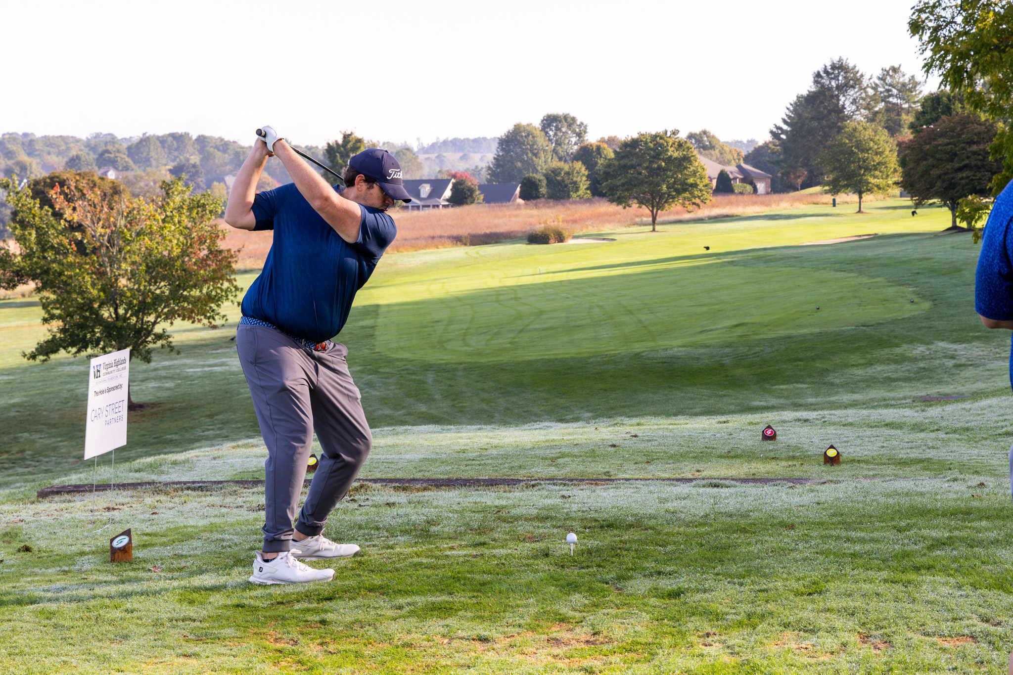 A golfer wearing a dark blue shirt and navy baseball cap mid-swing on a tee box during a sunny morning golf tournament, with open fairways, trees, and rolling hills in the background.
