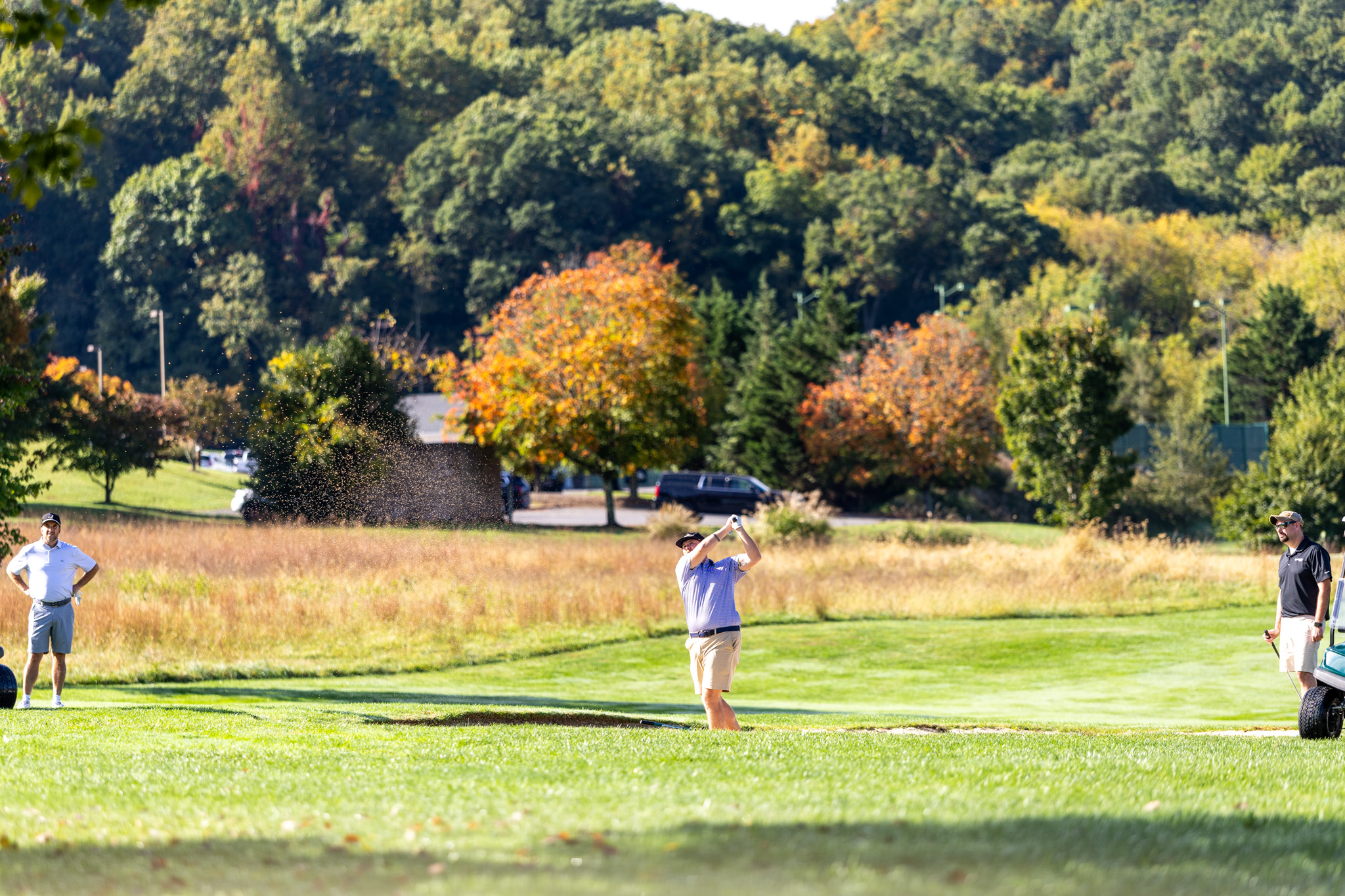 A golfer taking a shot out of a sand bunker on a bright, sunny day, with a sand spray in the air, framed by lush green lawns and vibrant autumn-colored trees on a hillside in the background.