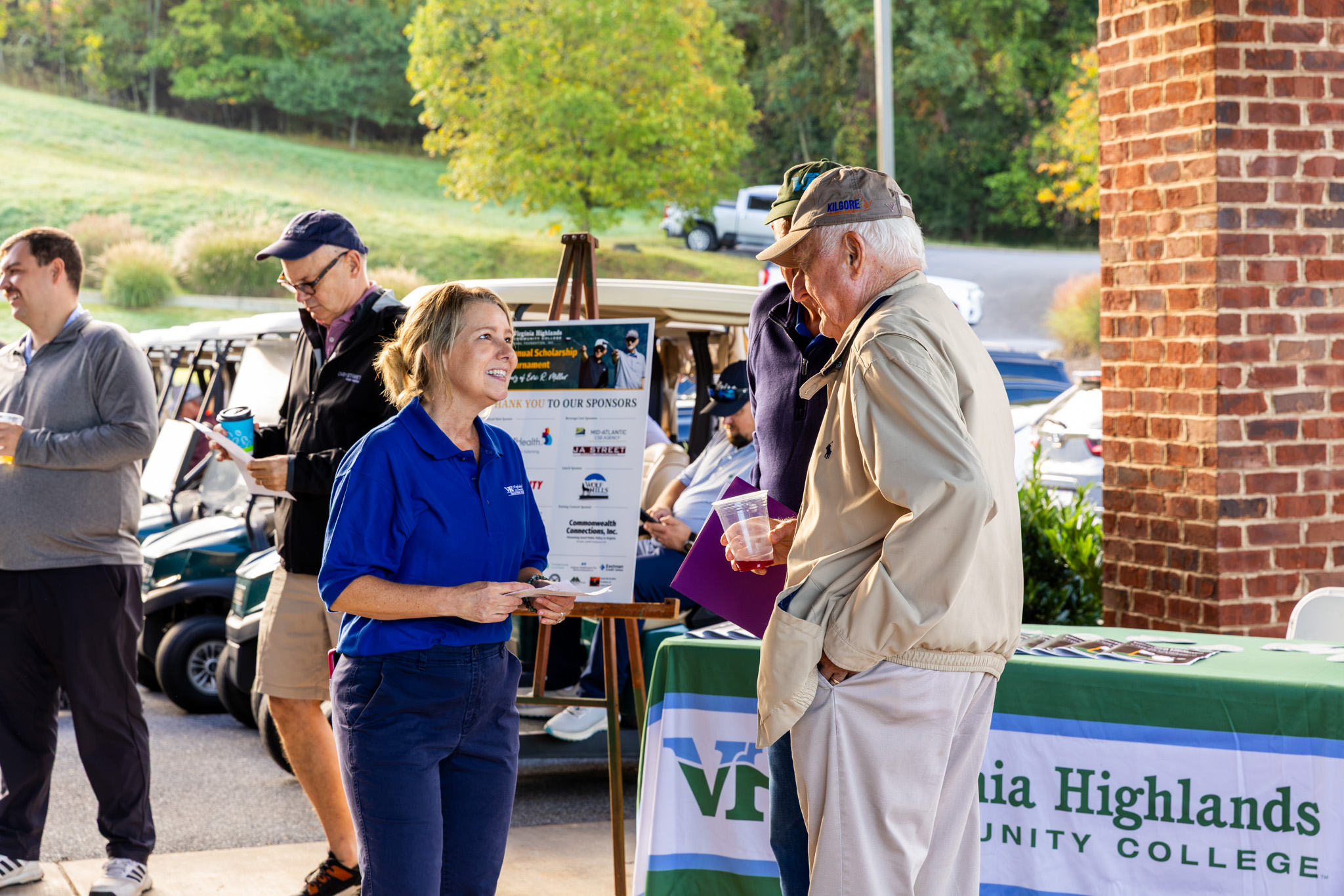 A woman wearing a blue Virginia Highlands Community College shirt talking with event attendees near a registration table decorated with a college banner and tournament sponsor sign.