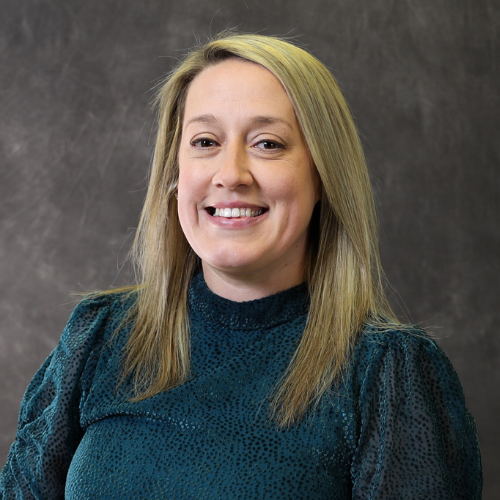 Portrait of Bethaney Hilt, a woman with long blonde hair, wearing a dark teal textured blouse against a gray studio background.