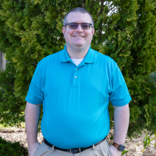 Brandon Hensley smiling outdoors in a bright blue polo shirt and glasses, standing in front of green shrubbery.