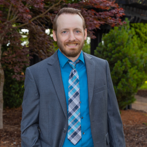 Derek Whisman, a man with a beard and light brown hair, wearing a grey suit jacket over a bright blue shirt and a patterned blue and grey tie.