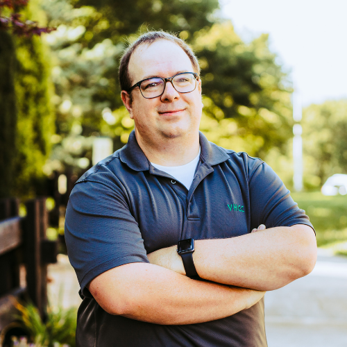 Josh Greenwell, smiling with arms crossed, wearing a grey polo shirt with a VHCC logo and glasses.