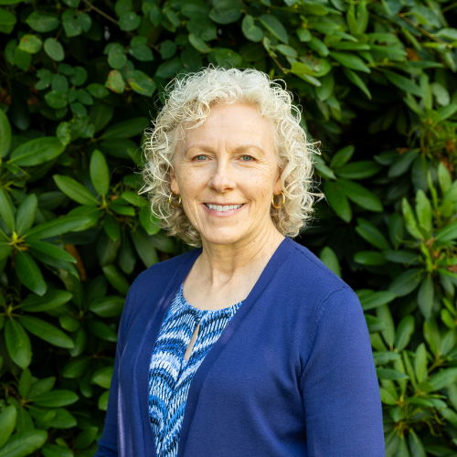 A smiling woman with curly, short blonde hair and hoop earrings, wearing a blue cardigan over a blue patterned blouse. She is standing outdoors in front of a dense background of green leafy bushes.