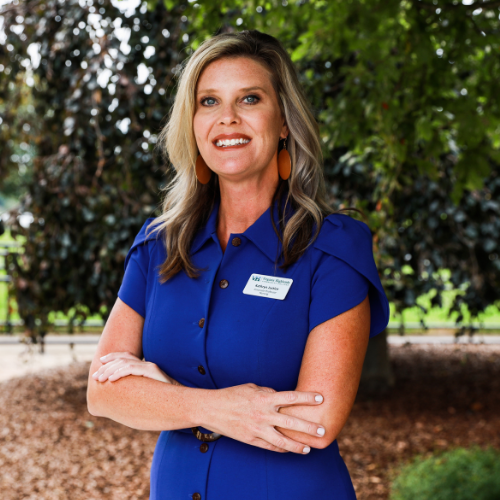 Portrait of Kathryn Justice, a woman with blonde hair, wearing a blue dress and large earrings, standing in front of lush green trees.