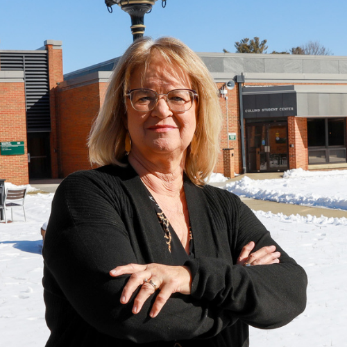 Headshot of Kathy Smith, a woman with blonde hair and glasses, posing with arms crossed. She is standing outdoors in a snowy courtyard in front of the Collins Student Center.