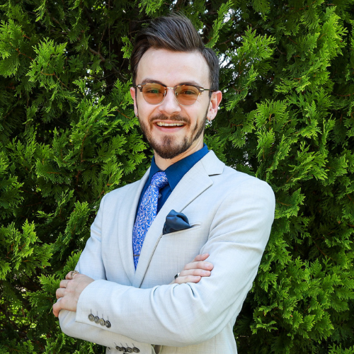 Ryan Whittington, a man with a beard and styled brown hair wearing a light grey suit, blue tie, and tinted glasses, standing with arms crossed against green hedges.