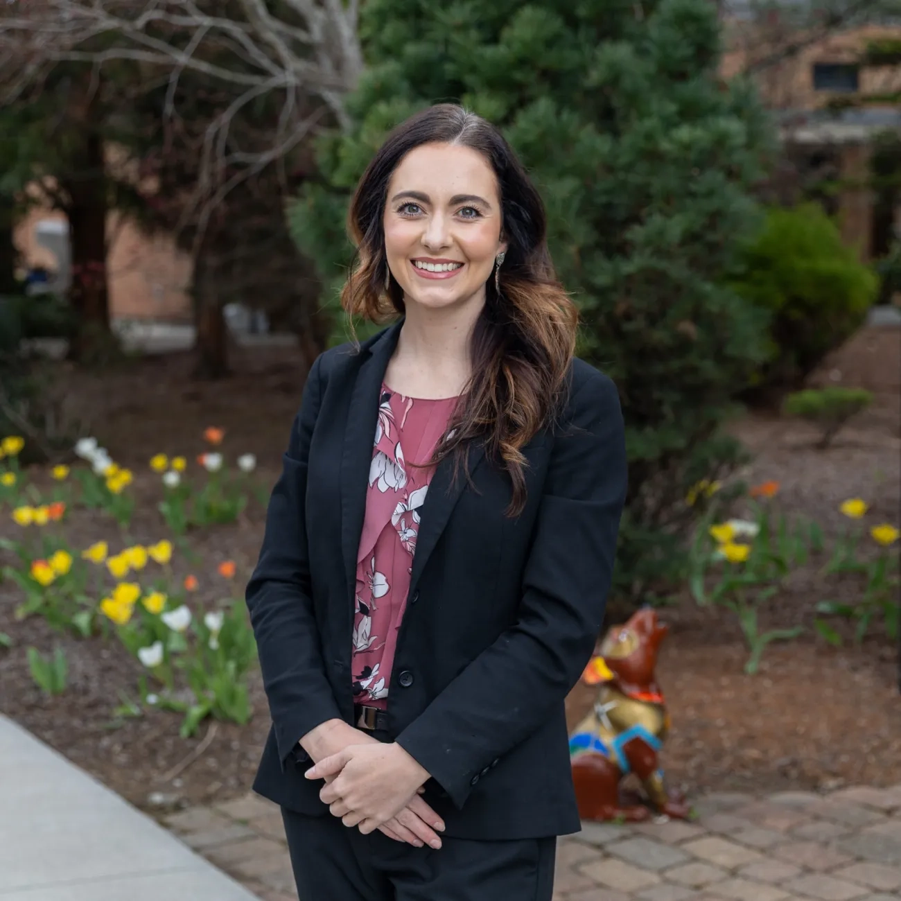 Professional outdoor portrait of VHCC alumna Leslie “Brooke” Rhymer smiling, wearing a black blazer and a pink floral blouse on a landscaped campus pathway.