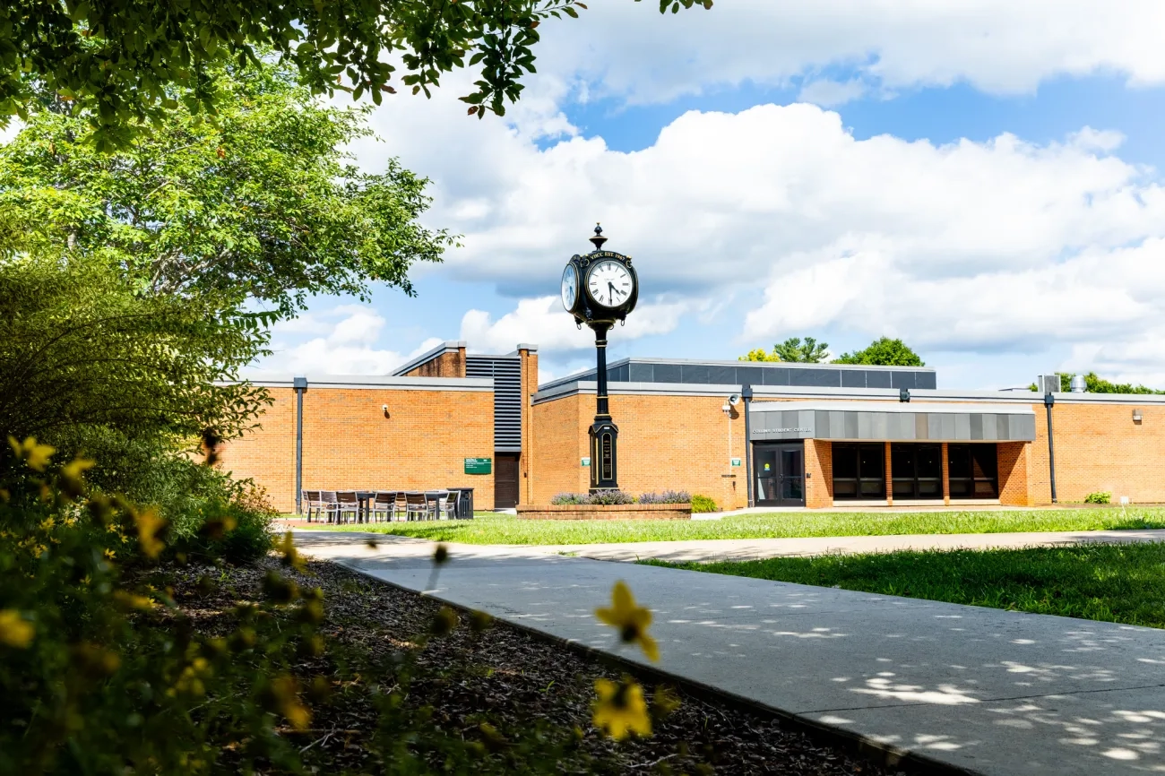 A scenic outdoor view of a brick campus building at Virginia Highlands Community College on a bright, sunny day. A tall, black traditional street clock stands in the courtyard under a blue sky with fluffy white clouds, framed by green foliage in the foreground.
