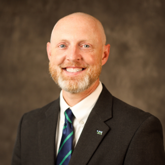 Headshot of Adam Hutchison, a man with a ginger beard and shaved head, wearing a dark suit, white shirt, and a blue and green striped tie.
