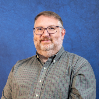 A professional headshot of a middle-aged man with short brown hair, a beard, and glasses, smiling and wearing a green checkered button-down shirt against a textured blue background.