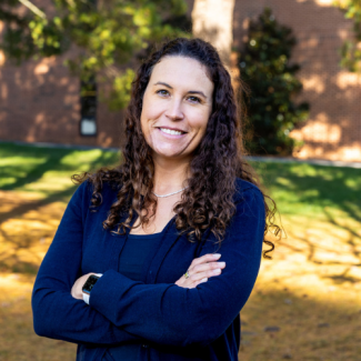 Alyssa Toney, a woman with long curly brown hair, wearing a dark blue cardigan over a black top, standing with arms crossed.