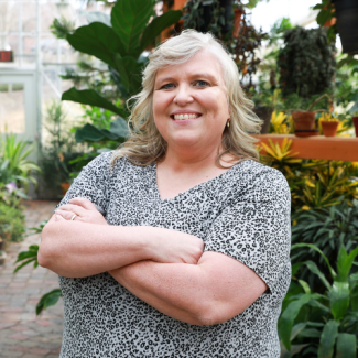 Headshot of Amy Baumgardener wearing a gray leopard print top, smiling with arms crossed in a bright greenhouse setting.