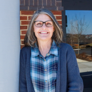 Andi Fultz wearing glasses and a blue plaid shirt under a gray cardigan, leaning against a white pillar with a brick building in the background.