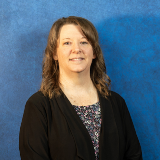 A professional headshot of Andrea Repass, a woman with shoulder-length wavy brown hair, smiling and wearing a black cardigan over a floral print blouse against a textured blue background.