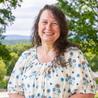 A smiling woman with long, curly brown hair, wearing a white blouse featuring a blue and orange floral pattern, standing outdoors with blurred green trees and mountains in the background.
