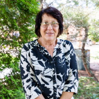 Beatrice Martino smiling in front of green foliage, wearing glasses and a black and white floral print button-down shirt.