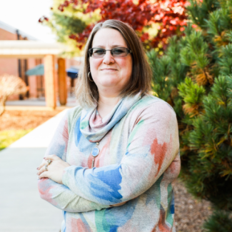 Becky Kell standing outdoors with arms crossed, wearing glasses and a colorful abstract-print sweater, with a walkway and autumn trees in the background.