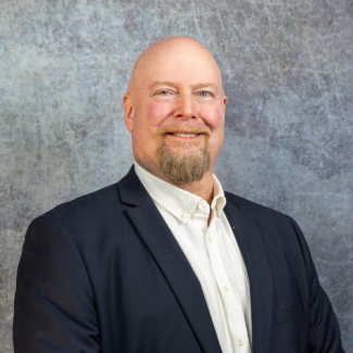 A smiling, bald man with a goatee and mustache, wearing a dark blue suit jacket over an unbuttoned white dress shirt, posing against a textured gray background.