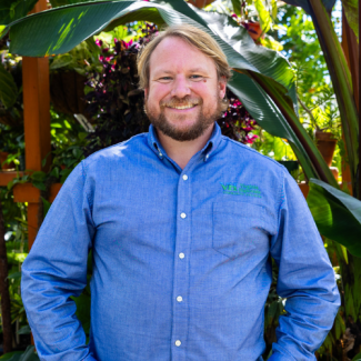 Headshot of Ben Casteel wearing a light blue button-down shirt, smiling in a greenhouse surrounded by large tropical plants.