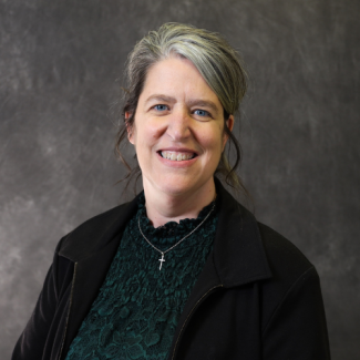 Headshot of Brigitte Casteel wearing a dark green lace top and black jacket, smiling against a neutral gray background.