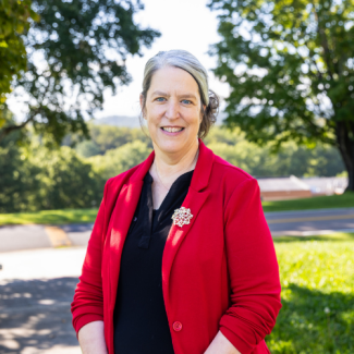 Headshot of Brigitte Casteel, a smiling woman with grey hair pulled back, wearing a bright red blazer with a brooch over a dark top outdoors.
