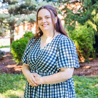 Headshot of Caitlin Manuel, a woman with long brown hair wearing a blue and white checkered dress, standing in a garden setting with her hands clasped.