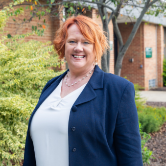 Headshot of Cheri Long, a woman with short, bright red hair wearing a white blouse and a navy blue blazer, smiling in an outdoor setting.