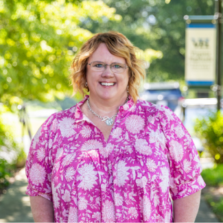 Cheri Long smiling outdoors, wearing glasses, a bright pink blouse with a white floral pattern, and a silver necklace with a round pendant, with green trees and a blurred campus sign in the background.