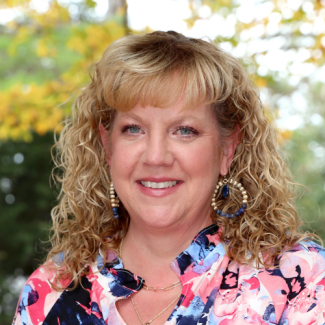 Headshot of Christine Fields, a woman with long, curly blonde hair and bangs wearing large hoop earrings and a pink, blue, and black floral blouse.
