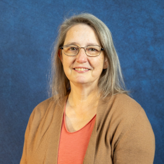 Professional headshot of Cindy Woosley, a woman with glasses and long grey hair, smiling and wearing a tan cardigan over a coral shirt against a blue background.