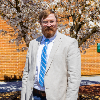 Headshot of Cory Lewis, a man with glasses and a red beard wearing a tan blazer and a blue striped tie, standing in front of a white flowering tree.
