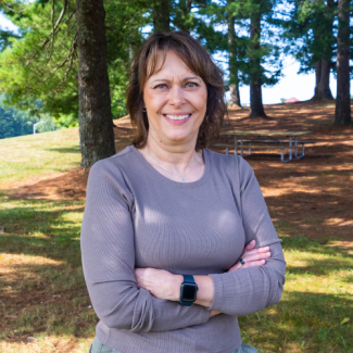 Headshot of D'Lisa Mullins, a woman with shoulder-length brown hair and bangs wearing a taupe long-sleeve shirt, standing outdoors with her arms crossed.