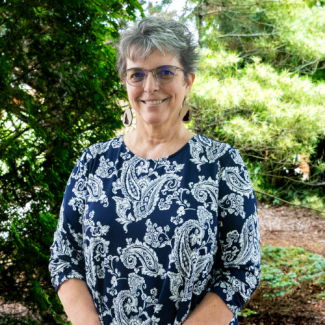 Headshot of Debbie Aylor wearing glasses and a navy blue paisley print top, smiling in front of lush garden greenery.