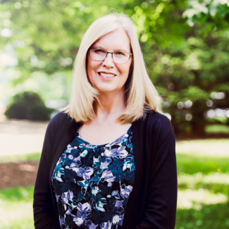 Deborah McCroskey, smiling outdoors, wearing a black cardigan over a floral print top and glasses.