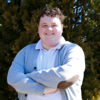 Headshot of Dillon Scott, a man with curly dark hair, smiling with arms crossed in a white polo and grey cardigan. He is standing outdoors in front of an evergreen tree.