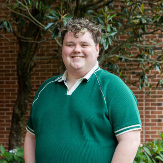 A smiling young man with curly, light brown hair, wearing a green polo shirt with white trim. He is standing outdoors in front of a brick wall and a leafy tree.