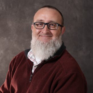 Donnie Melvin, smiling with a large grey beard and glasses, wearing a maroon fleece over a plaid shirt.