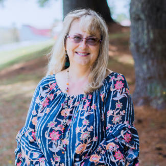 Headshot of Ella Ratcliffe-Sutherland, a woman with blonde hair and glasses, smiling in a floral patterned navy blouse. She is standing outdoors in a shaded area with trees.