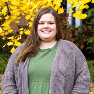 Headshot of Emileigh Yates, a woman with long brown hair, smiling in a green top and grey cardigan. She is standing outdoors in front of a tree with yellow autumn leaves.