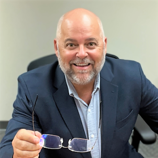 Ken Fairbanks, a man with a bald head and a graying beard, smiling while wearing a navy blazer and light blue shirt, holding a pair of eyeglasses toward the camera.