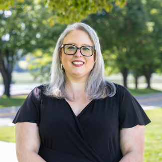 Headshot of Tiffanie Bishop, a woman with shoulder-length silver-grey hair and black-framed glasses, smiling outdoors in front of leafy green trees.