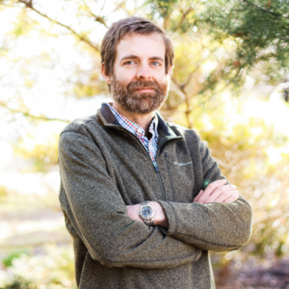 Jason Lachniet, smiling with a beard and arms crossed, wearing a brown quarter-zip sweater over a plaid shirt.