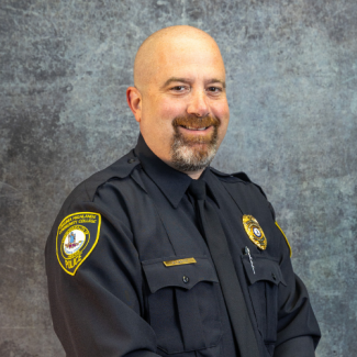 Professional headshot of John Carty, a man in a Virginia Highlands Community College Police uniform, smiling against a textured grey background.