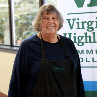 Portrait of Joyce Horn, a woman with short grey hair, wearing a navy shirt and a black apron emblazoned with the VHCC logo.