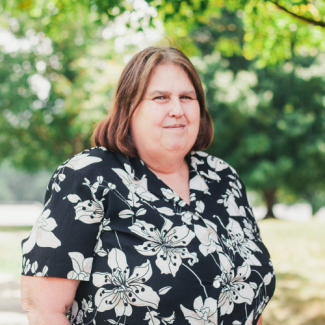 Portrait of Judy Miller, a woman with shoulder-length brown hair, wearing a black and white floral shirt outdoors.